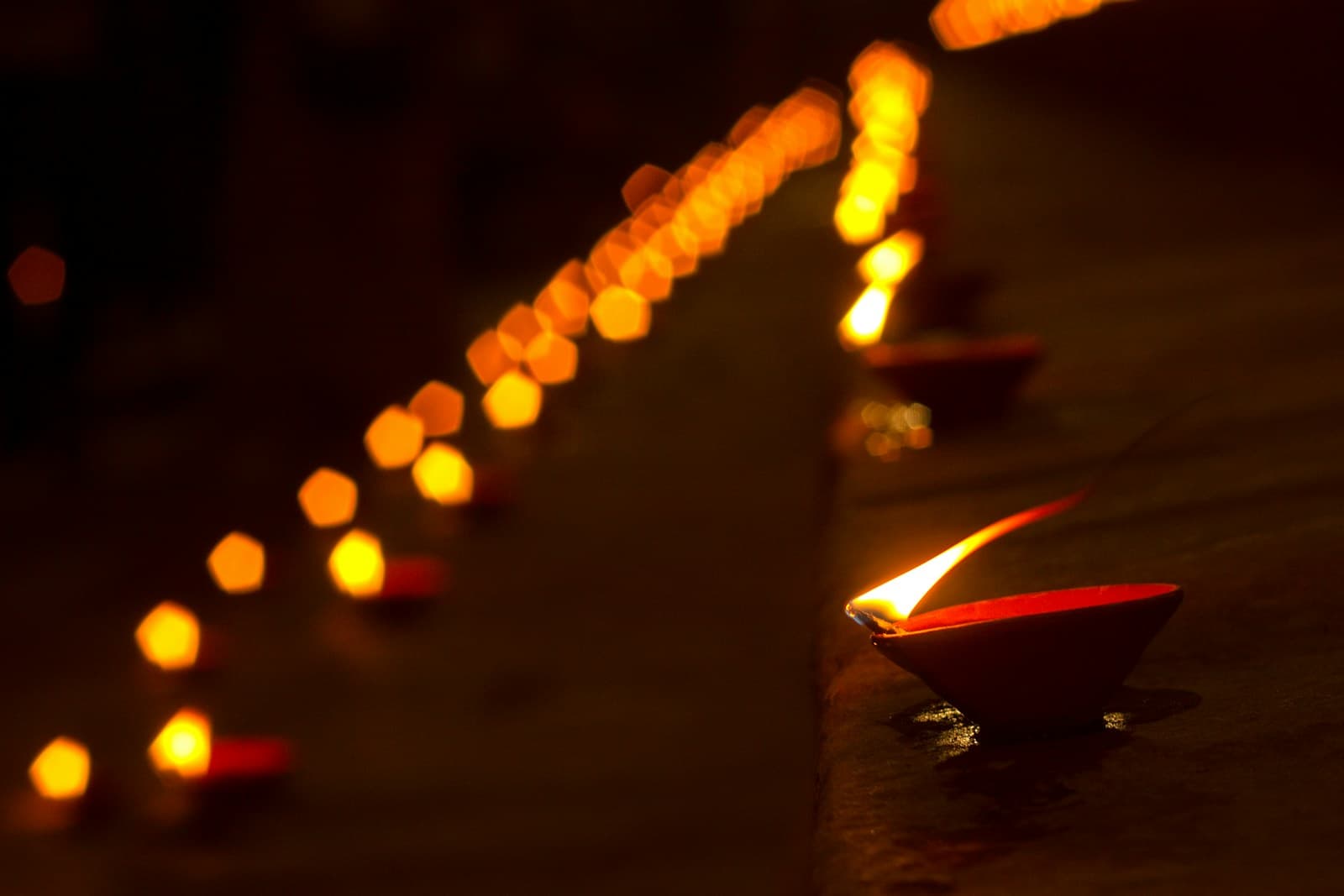 A single glowing diya on the ghat steps with a soft row of evening lamps in the background